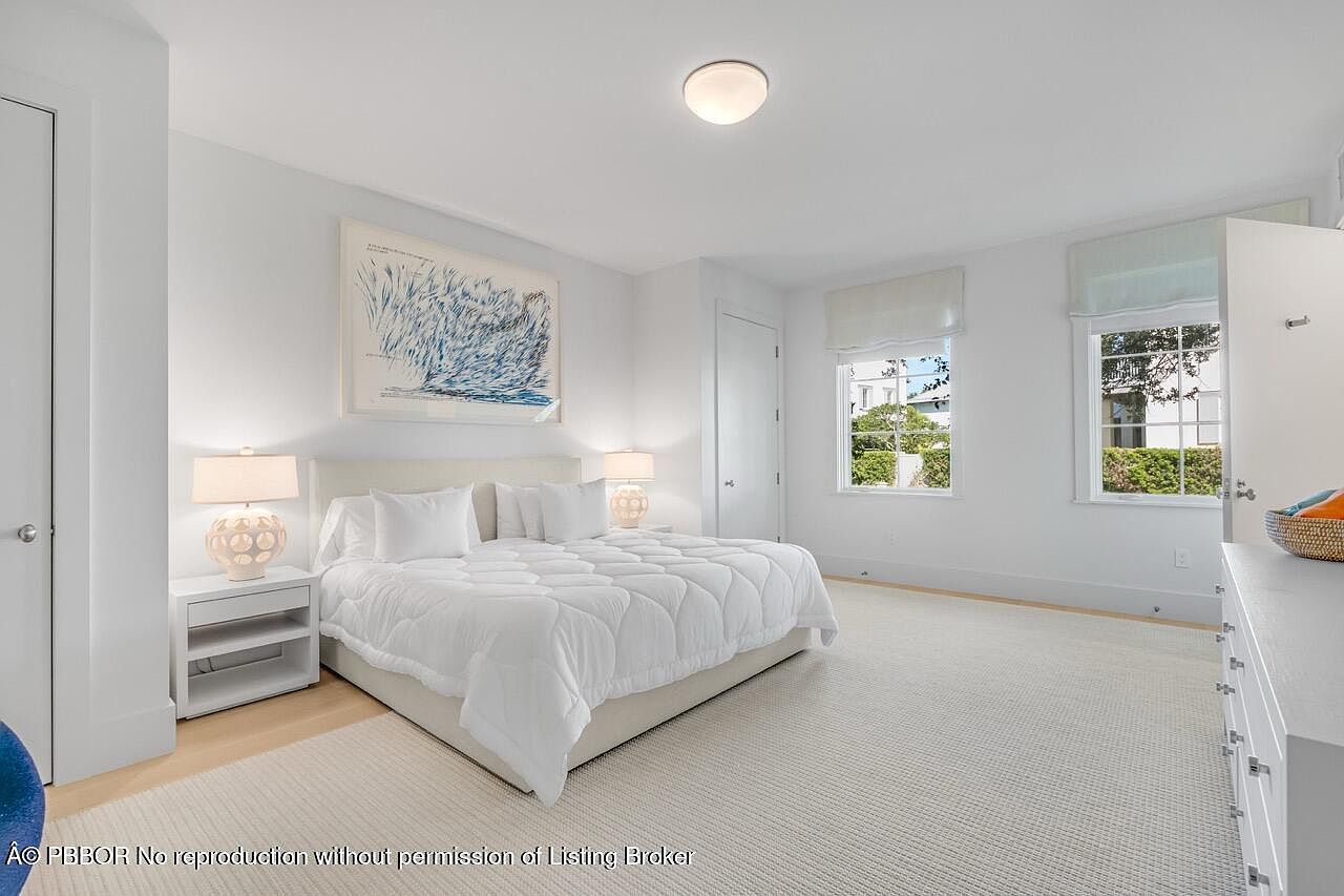 This is a bright and airy primary bedroom featuring a large bed with white linens, flanked by matching nightstands and lamps. A large piece of artwork hangs above the bed, and natural light streams in through two windows with white blinds. The room is carpeted and has a clean, minimalist aesthetic, creating a serene and inviting atmosphere.