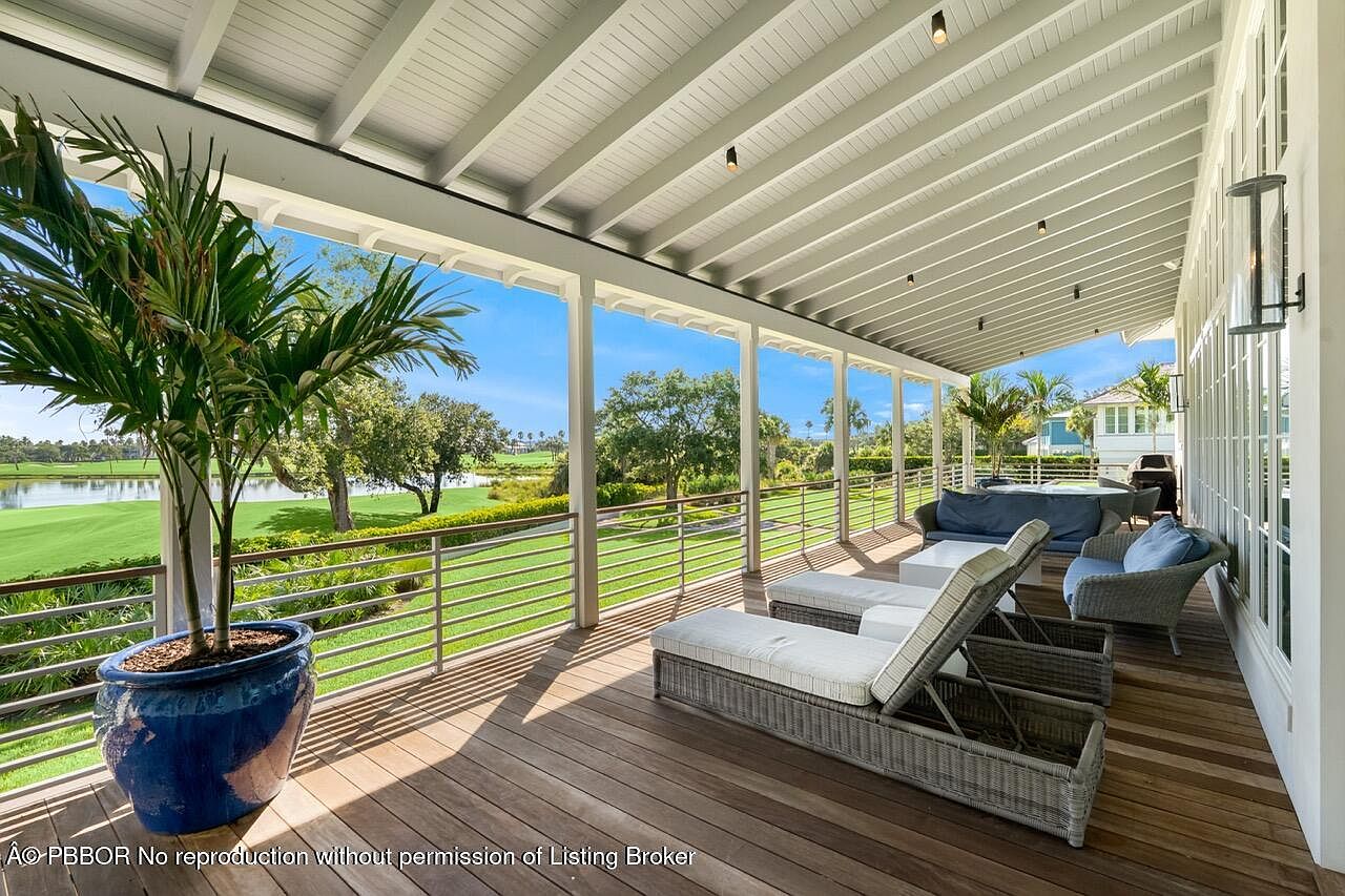 This image showcases a luxurious outdoor patio or deck area, featuring elegant lounge furniture and a potted palm tree. The space overlooks a beautifully manicured golf course, providing a serene and picturesque view. The wooden deck and white-painted ceiling create a sophisticated and inviting atmosphere, perfect for relaxation and entertainment.