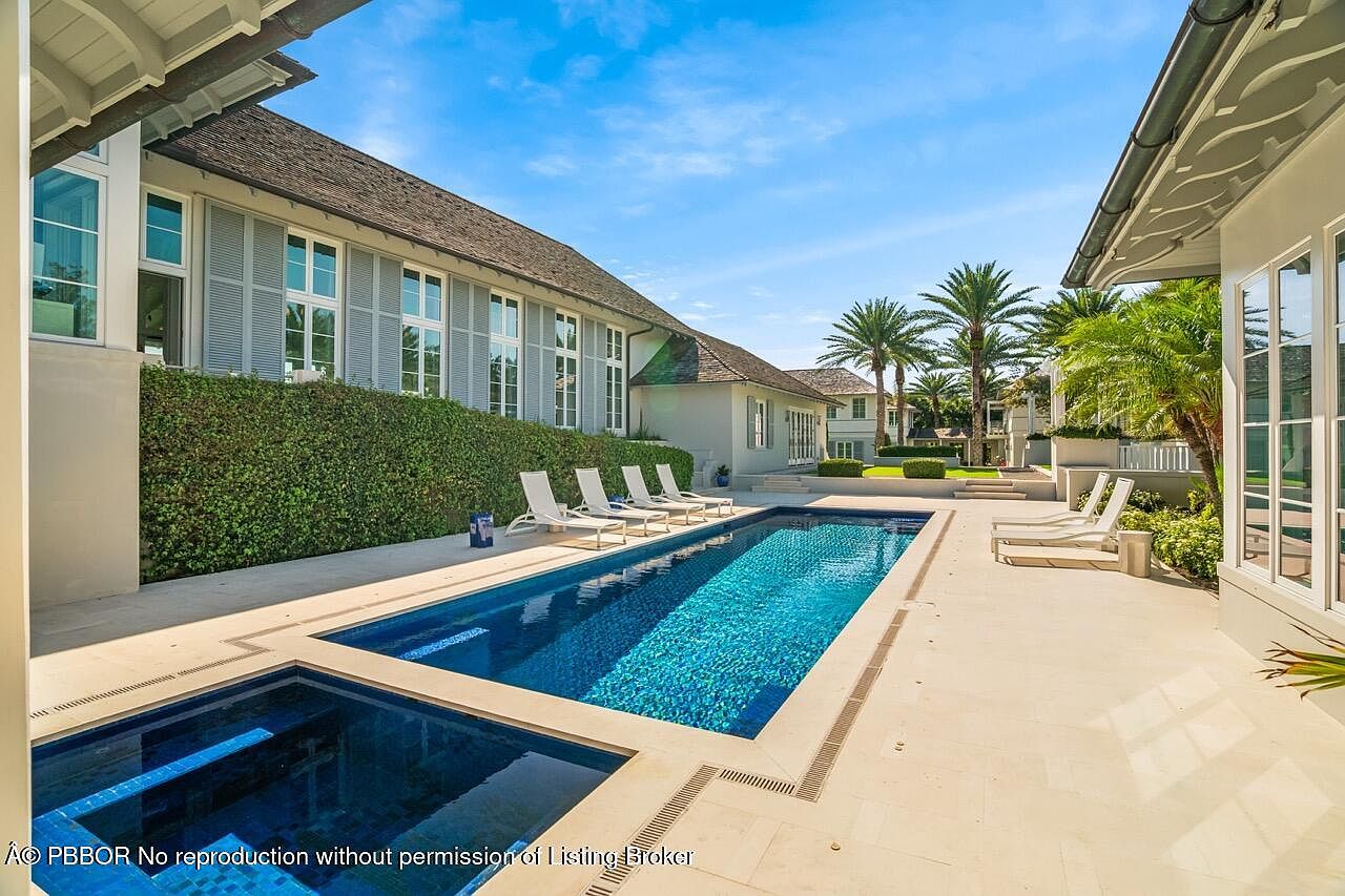 This image showcases a luxurious backyard pool and spa area. The rectangular pool features a dark blue tile interior, contrasting with the light-colored stone patio. Several lounge chairs are arranged around the pool, inviting relaxation, while lush greenery and palm trees add to the tropical ambiance. The architecture of the house is visible in the background, suggesting a high-end property.