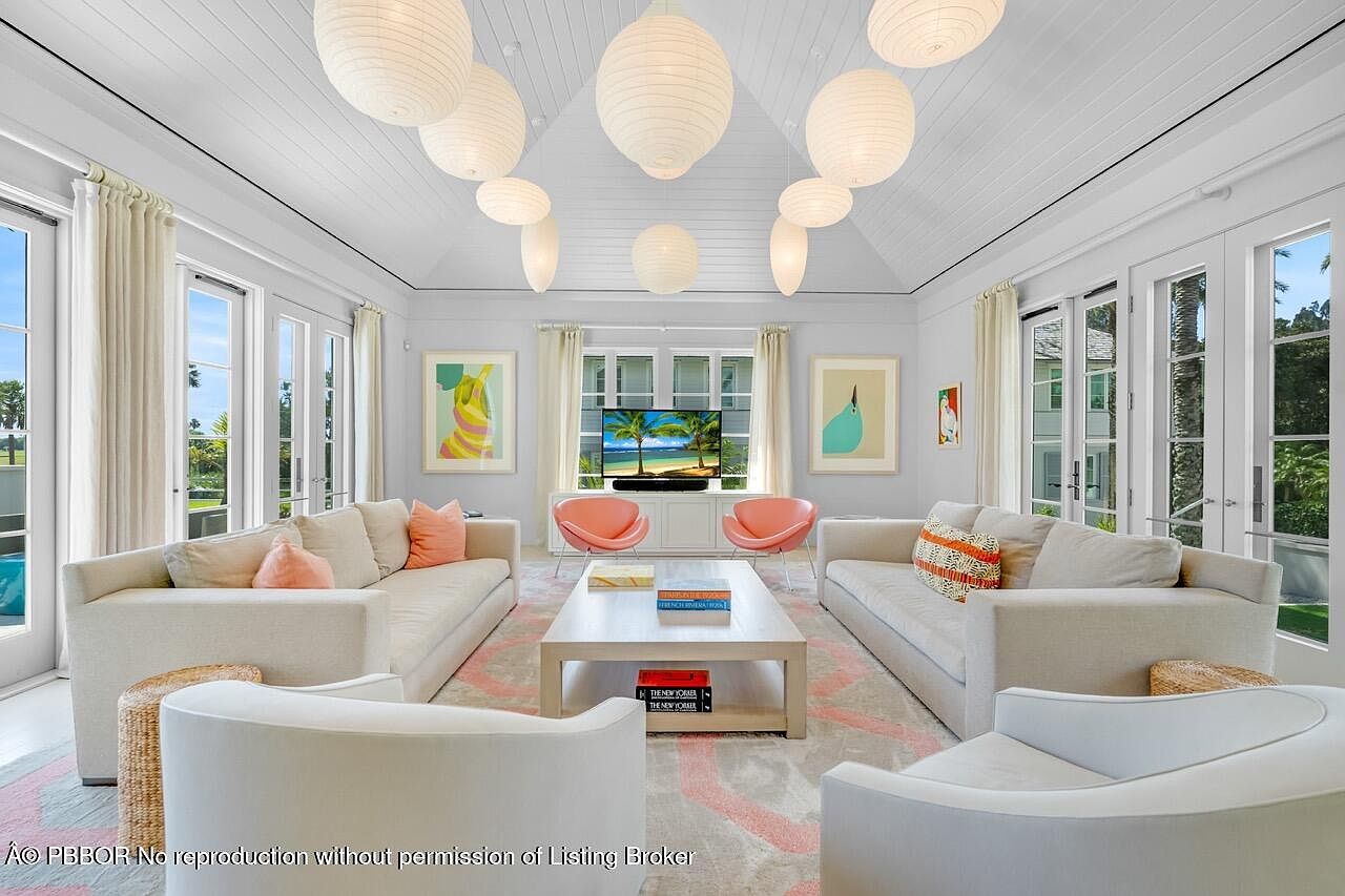 This is a bright and airy living room featuring two large sofas, two armchairs, and a light-colored rug with a subtle pattern. The room is illuminated by natural light streaming through multiple windows and glass doors, as well as several spherical pendant lights hanging from the vaulted ceiling. Artwork adorns the walls, and the overall aesthetic is clean, modern, and inviting.