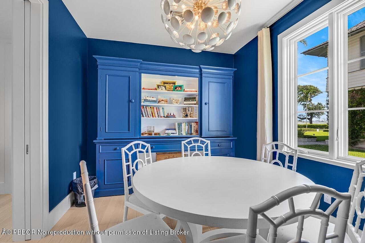 This is an interior shot of a dining room featuring a round white table surrounded by white chairs with a unique design. A large blue hutch with open shelving and closed cabinets stands against the blue wall, and a modern chandelier hangs from the ceiling. A window provides natural light and a view of the outdoors.