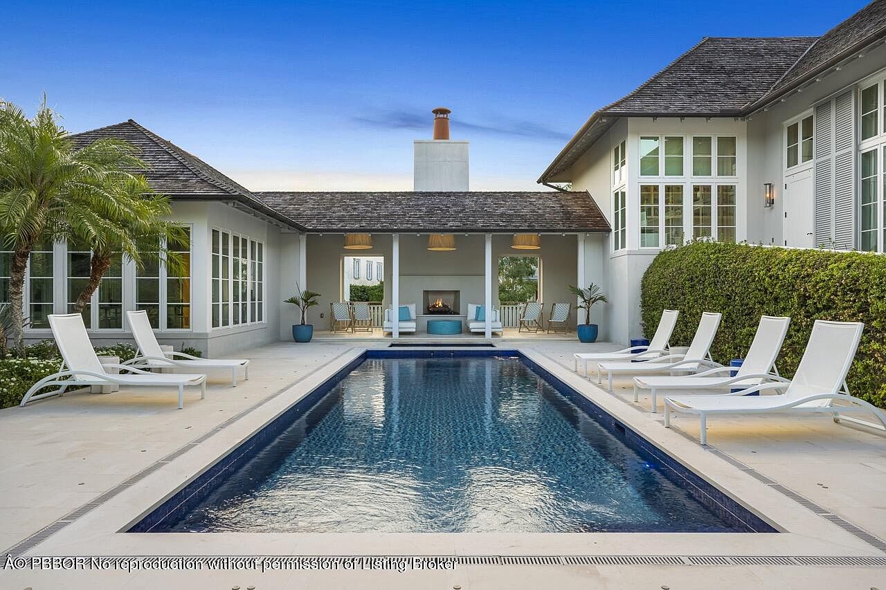 This is an exterior shot of a luxurious pool area. The rectangular pool is surrounded by white lounge chairs and lush greenery. A covered outdoor living space with a fireplace and seating area is visible in the background, creating an inviting and relaxing atmosphere.