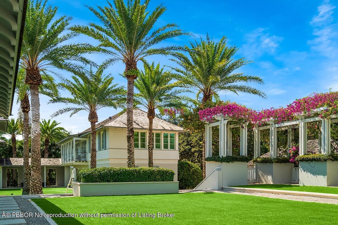 This image showcases a beautifully landscaped yard featuring lush green grass, mature palm trees, and a charming pergola adorned with vibrant pink flowers. The property includes a well-maintained building with a light-colored exterior and a neatly trimmed hedge, creating an inviting and serene outdoor space. The clear blue sky enhances the overall appeal of this picturesque setting.