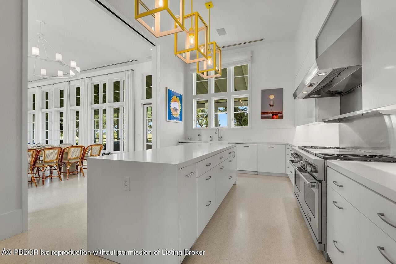 This is a bright and spacious kitchen featuring a large white island with drawers and cabinets, stainless steel appliances, and modern yellow pendant lighting. The kitchen opens to a dining area with large windows and white curtains, creating a seamless transition between spaces. The overall impression is clean, modern, and inviting.