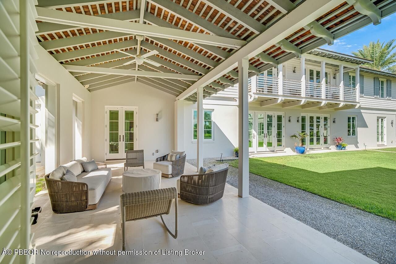This image showcases a beautifully designed outdoor patio area, featuring comfortable wicker furniture arranged on a light-colored stone patio. The patio is covered by a stylish roof structure with exposed beams, providing shade and architectural interest. The space seamlessly transitions to a well-manicured lawn and gravel path, with the main house visible in the background, complete with balconies and French doors, creating an inviting indoor-outdoor living experience.