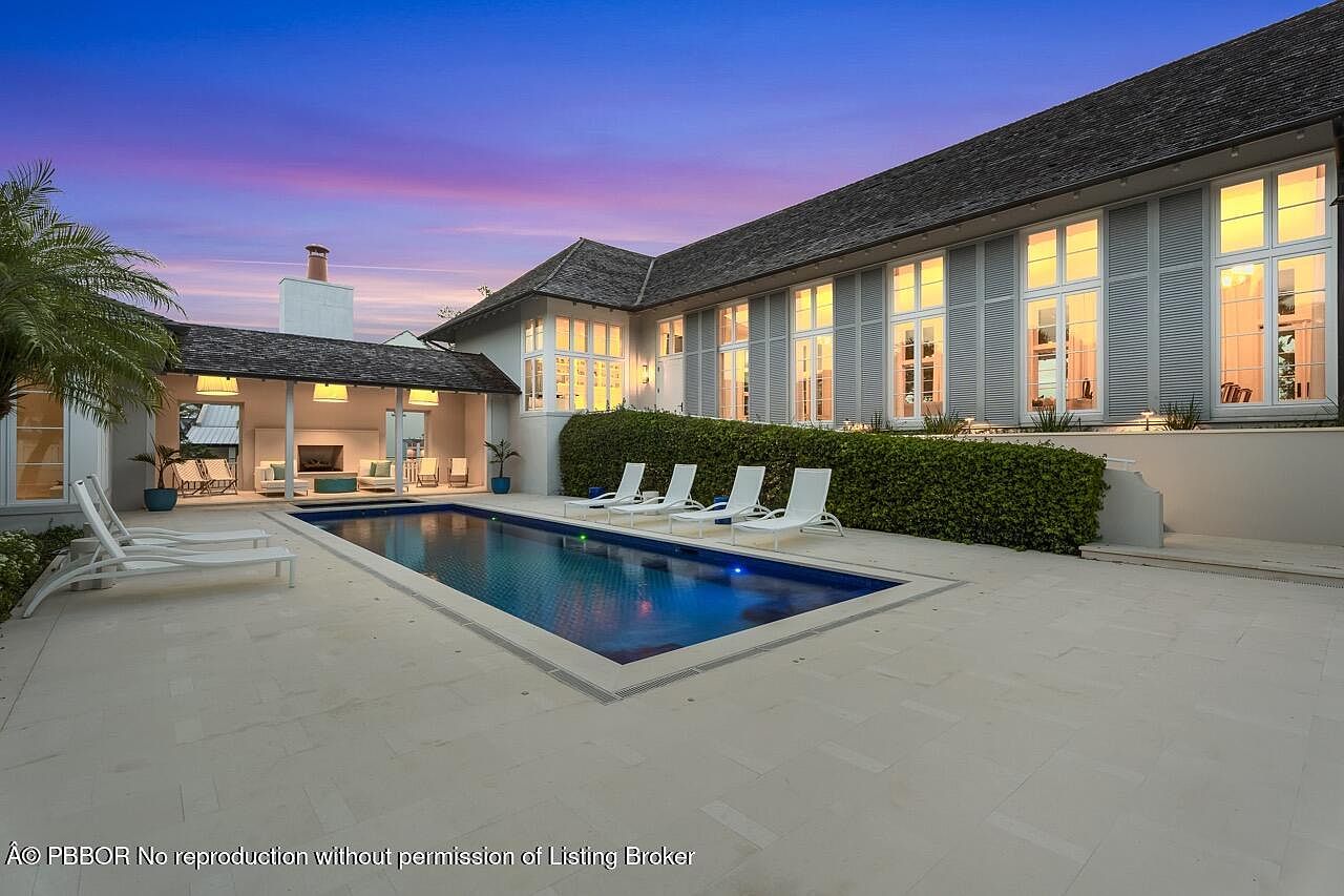 This is an exterior shot of a luxurious home featuring a rectangular pool with blue water, surrounded by a light-colored stone patio. Several white lounge chairs are arranged around the pool, and a well-maintained hedge separates the pool area from the house. The house itself has large windows with shutters and a dark roof, creating a sophisticated and inviting atmosphere.