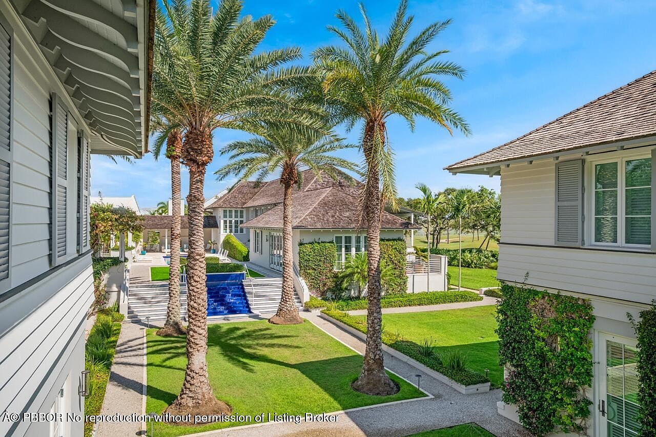 This image showcases the rear exterior of a luxurious home, featuring a well-manicured lawn, a blue-tiled water feature, and several palm trees that add a tropical touch. The architecture includes white siding, grey shutters, and a brown shingle roof, creating a sophisticated and inviting atmosphere. The view is from an elevated position, providing a comprehensive look at the property's backyard and landscaping.
