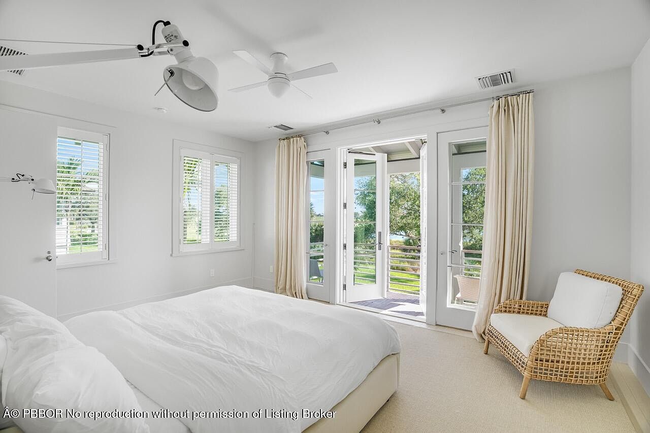 This is a bright and airy primary bedroom featuring a large bed with white linens, two windows with white shutters, and French doors leading to a balcony. A wicker chair with white cushions sits in the corner, adding a touch of texture to the otherwise all-white color scheme. The room exudes a sense of calm and relaxation.