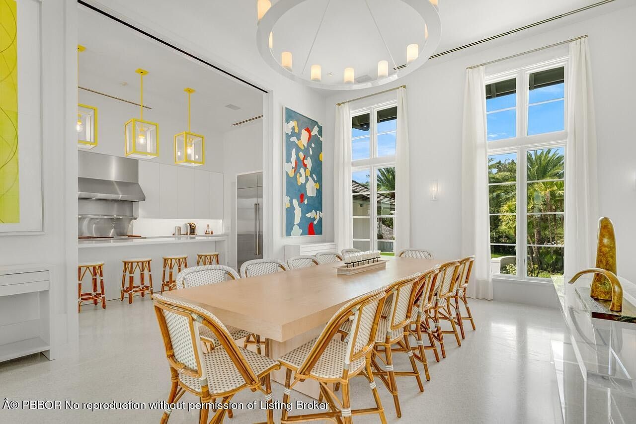 This is an interior shot of a dining room that flows into a kitchen area. The dining room features a long wooden table surrounded by wicker chairs, illuminated by a modern circular chandelier. Large windows with white curtains offer a view of the exterior, and an abstract painting adds a touch of color to the white walls.
