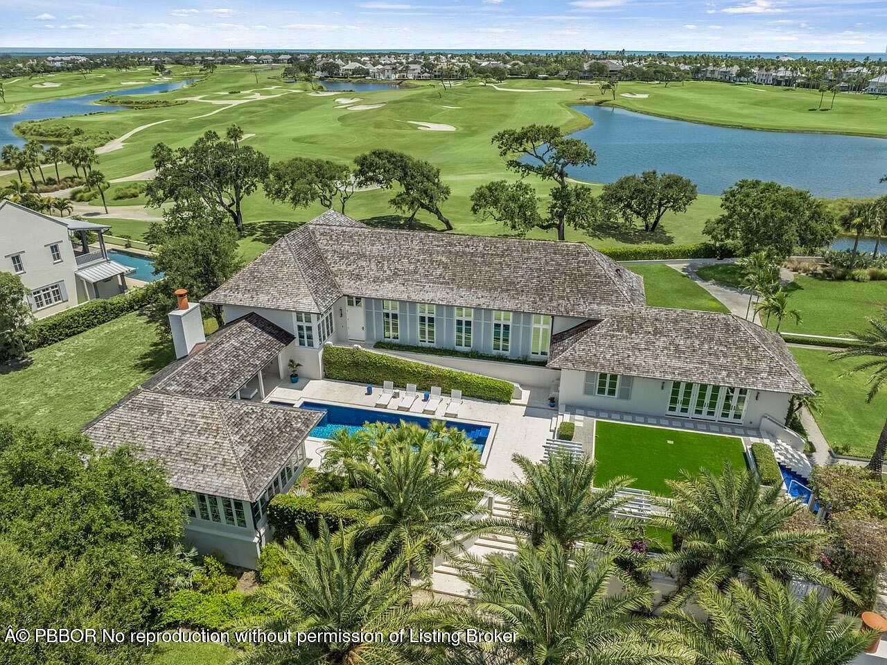 This aerial shot showcases a luxurious estate featuring a large, light-colored house with a gray roof, a swimming pool, and meticulously landscaped grounds with palm trees. The property backs onto a golf course with water features, offering stunning views and a sense of exclusivity. The overall impression is one of opulence and tranquility.