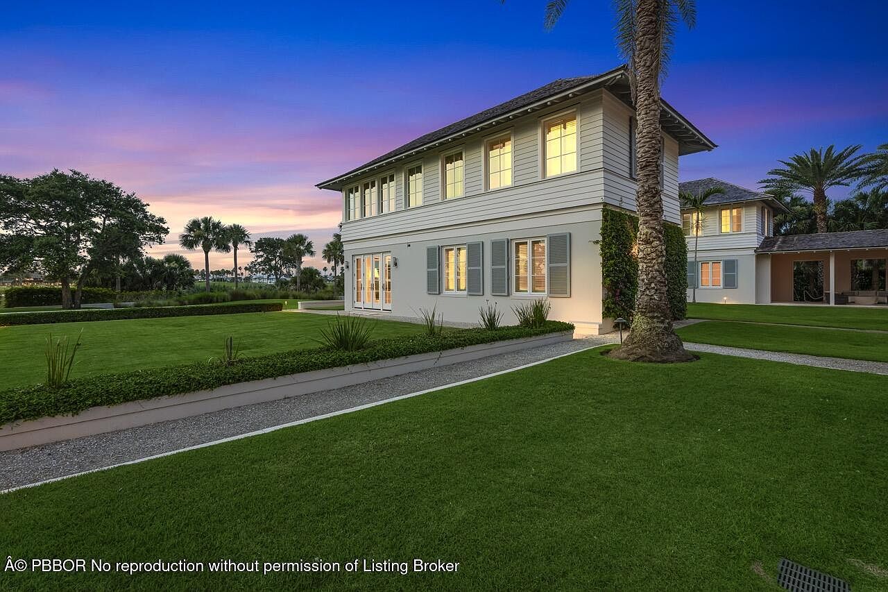 This image showcases the front exterior of a beautiful two-story home with a well-manicured lawn. The house features a light-colored facade, gray shutters, and a dark roof, exuding a classic and elegant style. A gravel pathway leads to the entrance, and lush greenery surrounds the property, enhancing its curb appeal and creating a serene atmosphere.