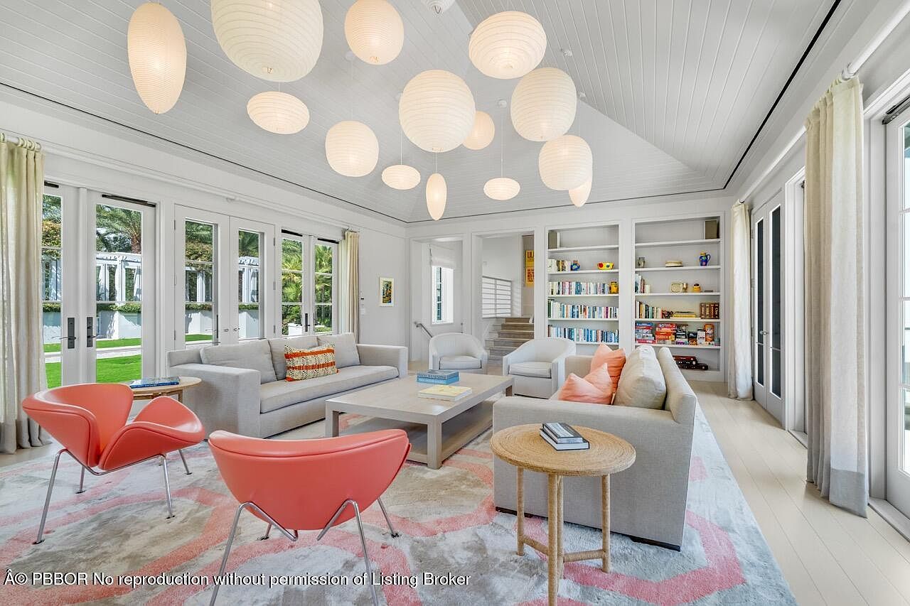This is an interior shot of a bright and airy living room. The room features a high vaulted ceiling adorned with multiple paper lanterns, creating a soft, diffused light. Two sofas, two coral-colored chairs, and a light-colored rug create a comfortable seating area, while built-in bookshelves add a touch of sophistication. French doors lead to the outside, enhancing the room's open and inviting atmosphere.