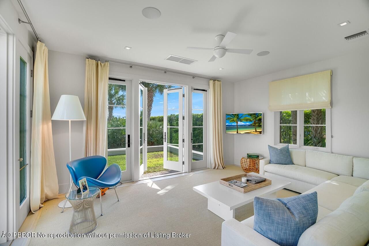 This is an interior shot of a bright and airy living room. The room features a white sectional sofa with blue pillows, a modern coffee table, and a blue accent chair. Large glass doors open to a lush green lawn, bringing the outdoors in and creating a seamless transition between indoor and outdoor living spaces.