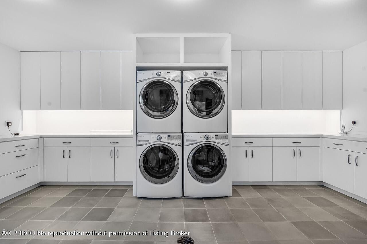 This is a well-organized laundry room featuring a stacked washer and dryer unit centered between white cabinetry. The countertops are also white, providing ample workspace, and under-cabinet lighting enhances the brightness. The floor is tiled in a neutral tone, contributing to the room's clean and modern aesthetic.