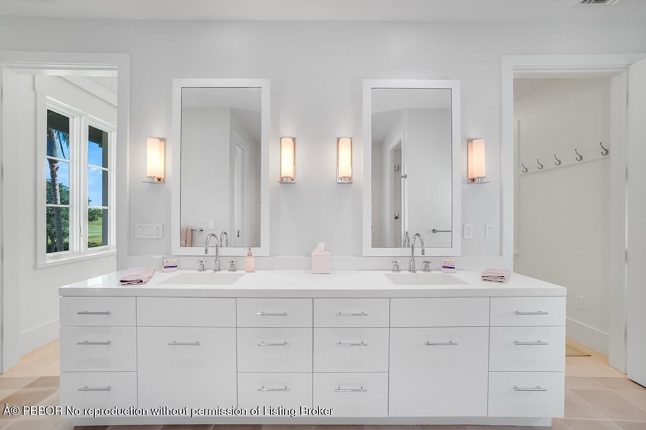 This is a bright and modern primary bathroom featuring a double vanity with white cabinetry and countertops. Two large mirrors are mounted above the sinks, each flanked by sconce lighting. The room has a clean and minimalist aesthetic, with neutral tones and ample storage space.