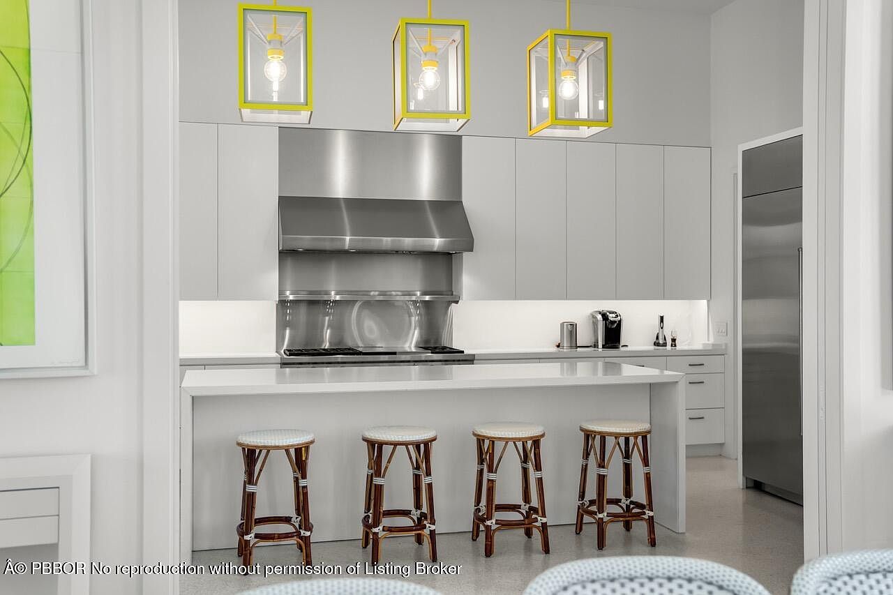 This is a bright and modern kitchen featuring sleek white cabinetry, stainless steel appliances, and a large island with a white countertop. Four bar stools with woven seats are positioned at the island, and three yellow pendant lights hang above. The kitchen has a clean and minimalist aesthetic.
