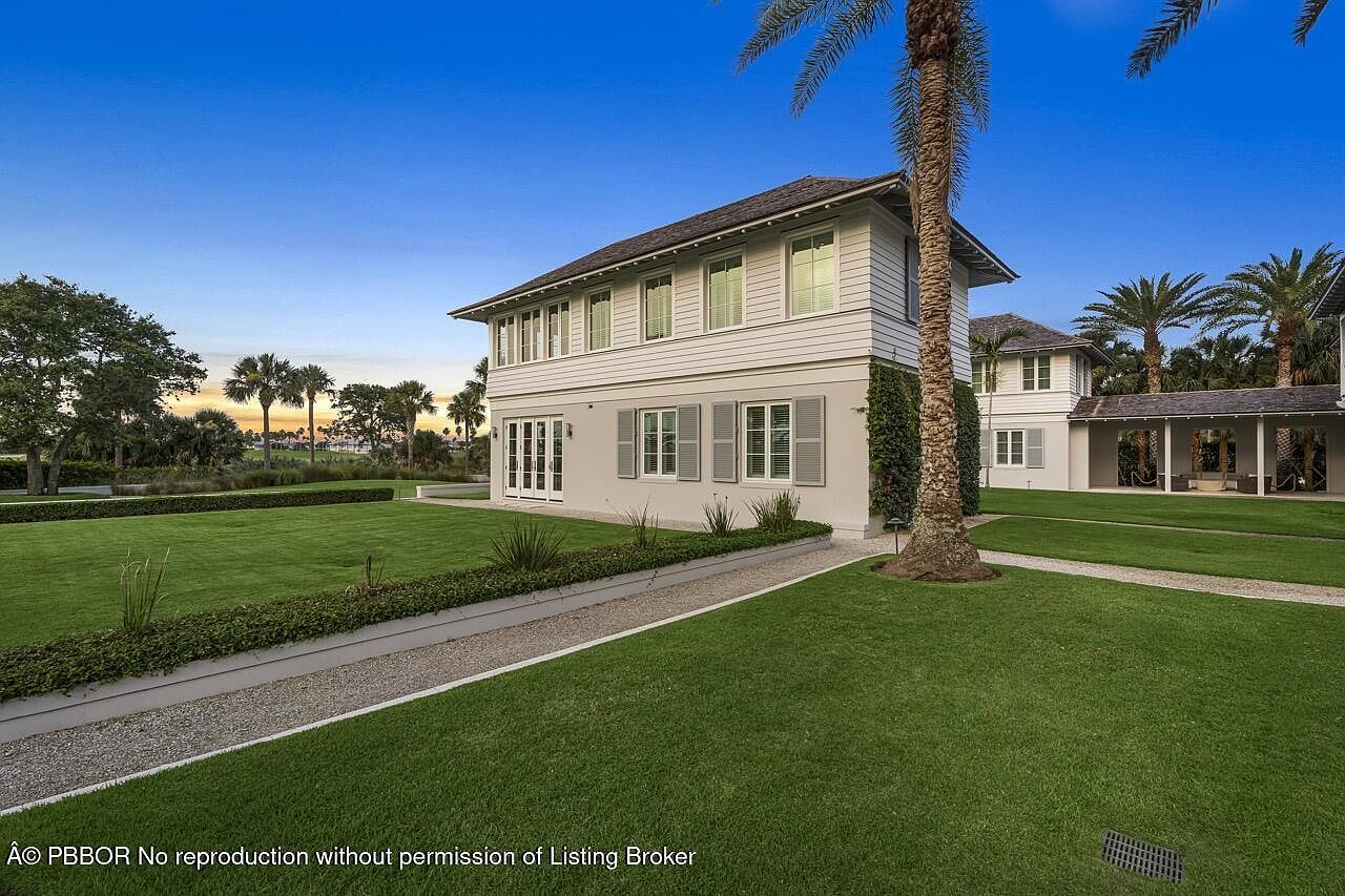 This is a front exterior view of a two-story house with a well-manicured lawn and landscaping. The house features a light-colored facade with gray shutters and a dark roof. A gravel pathway leads to the house, and palm trees are visible in the background, adding to the tropical feel of the property.