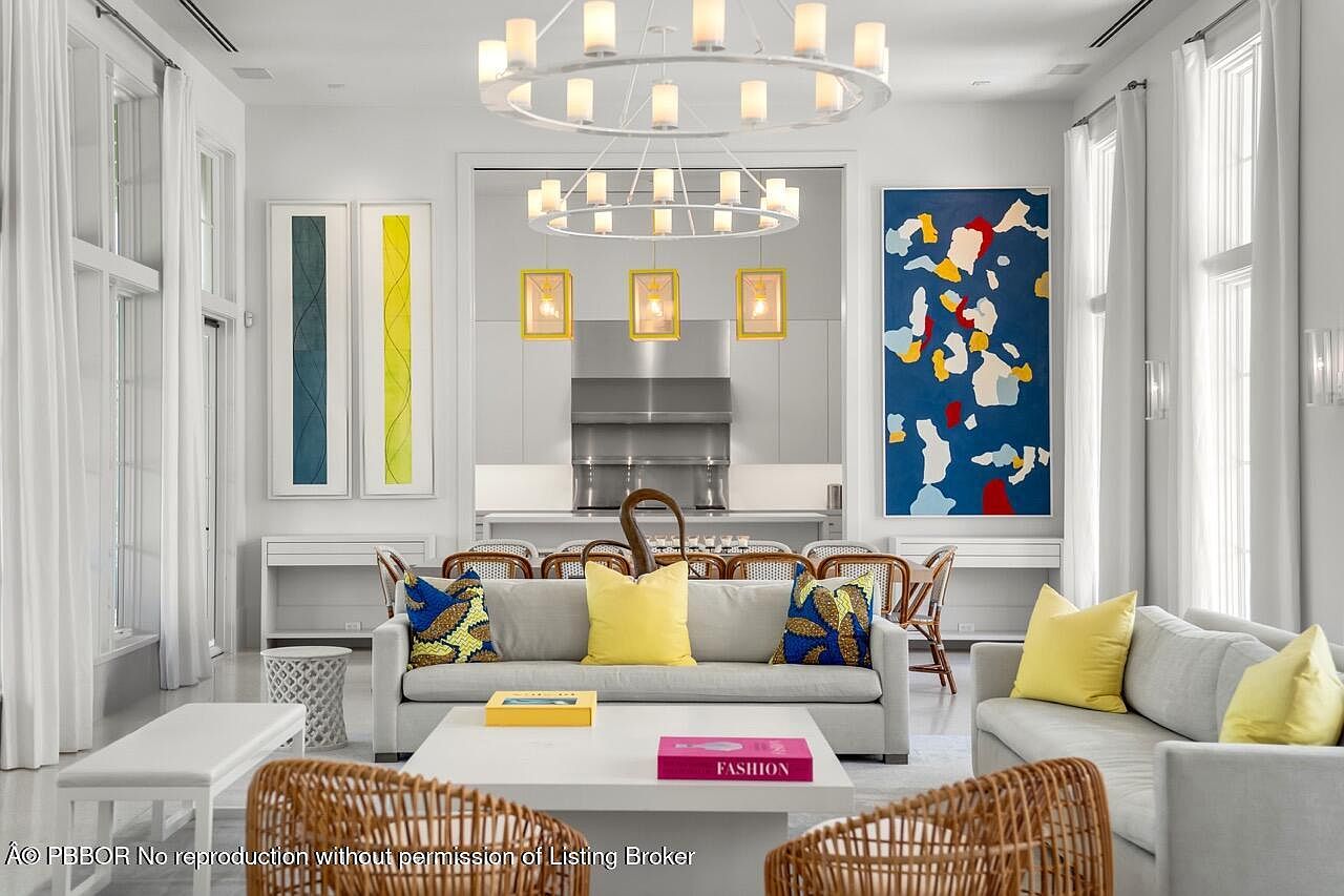 This is an interior shot of a modern living room that seamlessly transitions into a dining area and kitchen. The living room features two light gray sofas, wicker chairs, and a white coffee table with books. The room is brightly lit by natural light from large windows and an elegant chandelier, complemented by colorful artwork on the walls, creating a sophisticated and inviting atmosphere.