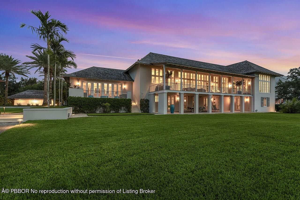 This is a front exterior view of a large, luxurious two-story home with a sprawling green lawn. The house features a covered balcony that spans most of the second floor, supported by columns. The architecture is elegant and inviting, with a well-maintained landscape that enhances the property's curb appeal, all under a beautiful sunset sky.