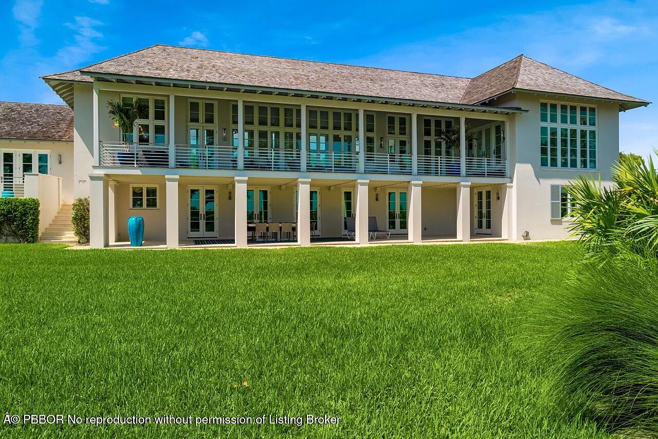 This is a front exterior view of a large, two-story house with a light-colored facade and a gray shingle roof. The house features a covered balcony on the second floor with multiple sets of French doors and a series of columns supporting the balcony. A well-manicured green lawn extends in front of the house, adding to its curb appeal.
