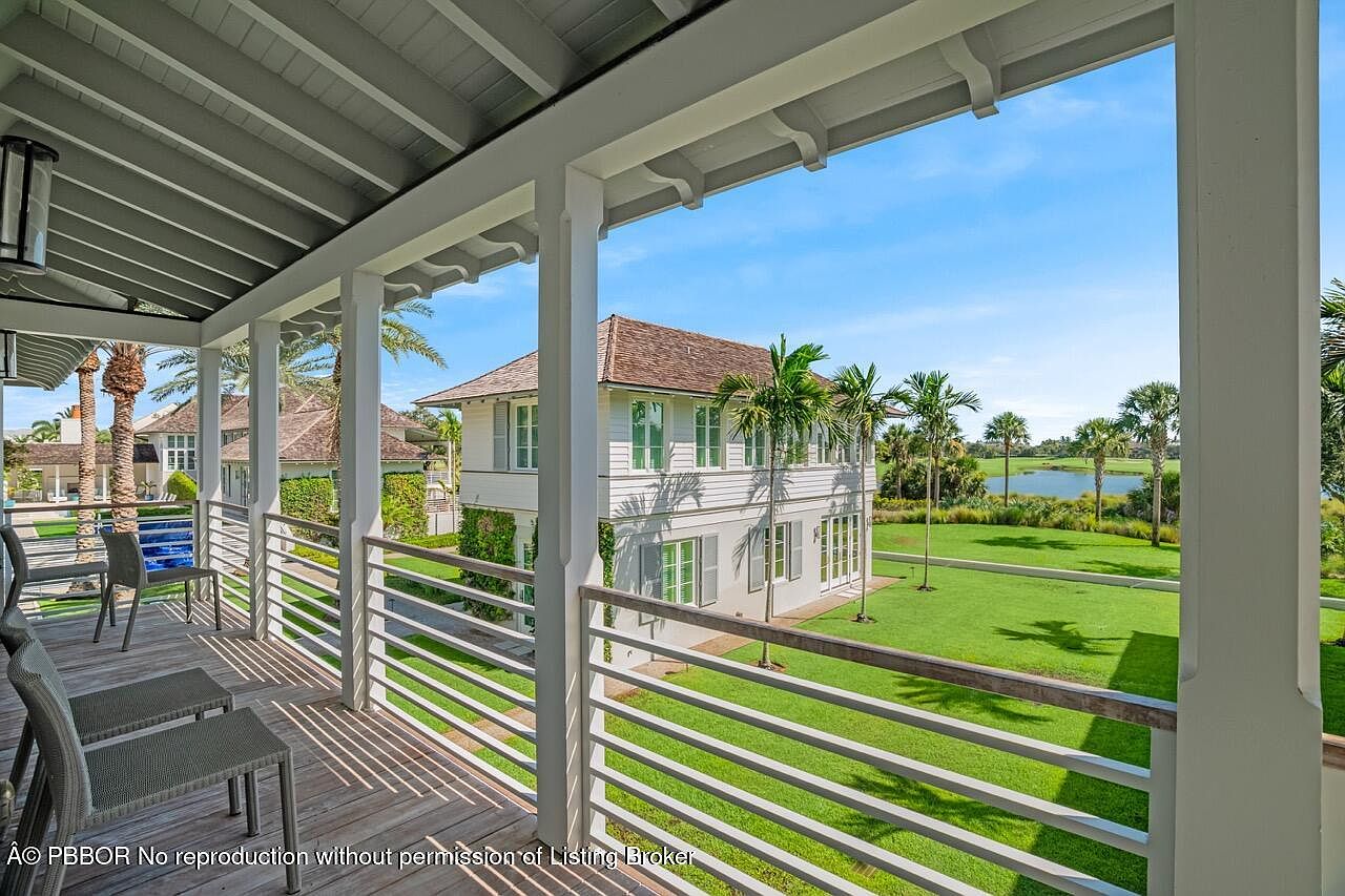 This image showcases a beautiful outdoor balcony or deck area with a view of a well-manicured lawn and a two-story house. The balcony features white railings and columns, providing a clean and elegant aesthetic. The house in the background has a brown roof and white exterior, complemented by lush greenery and palm trees, creating a serene and luxurious atmosphere.