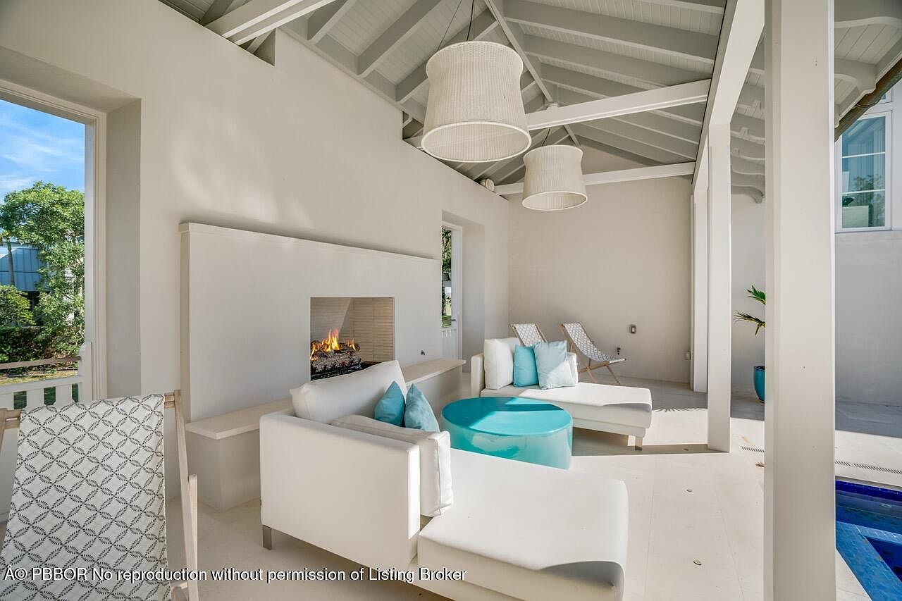 This image showcases a luxurious outdoor living space featuring a fireplace, comfortable white seating with blue accent pillows, and stylish pendant lighting. The area is designed with a clean, modern aesthetic, offering a relaxing and elegant atmosphere. The space transitions seamlessly to an outdoor pool area, enhancing the property's appeal for entertaining and leisure.