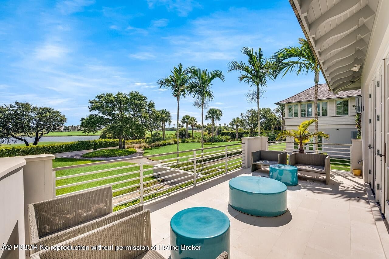 This image showcases a luxurious patio or balcony area with stylish outdoor furniture, including wicker chairs and vibrant blue ceramic tables. The space overlooks a beautifully manicured lawn, palm trees, and a glimpse of a nearby body of water, suggesting a high-end property with stunning views. The architecture of the adjacent building is elegant, contributing to the overall upscale ambiance.