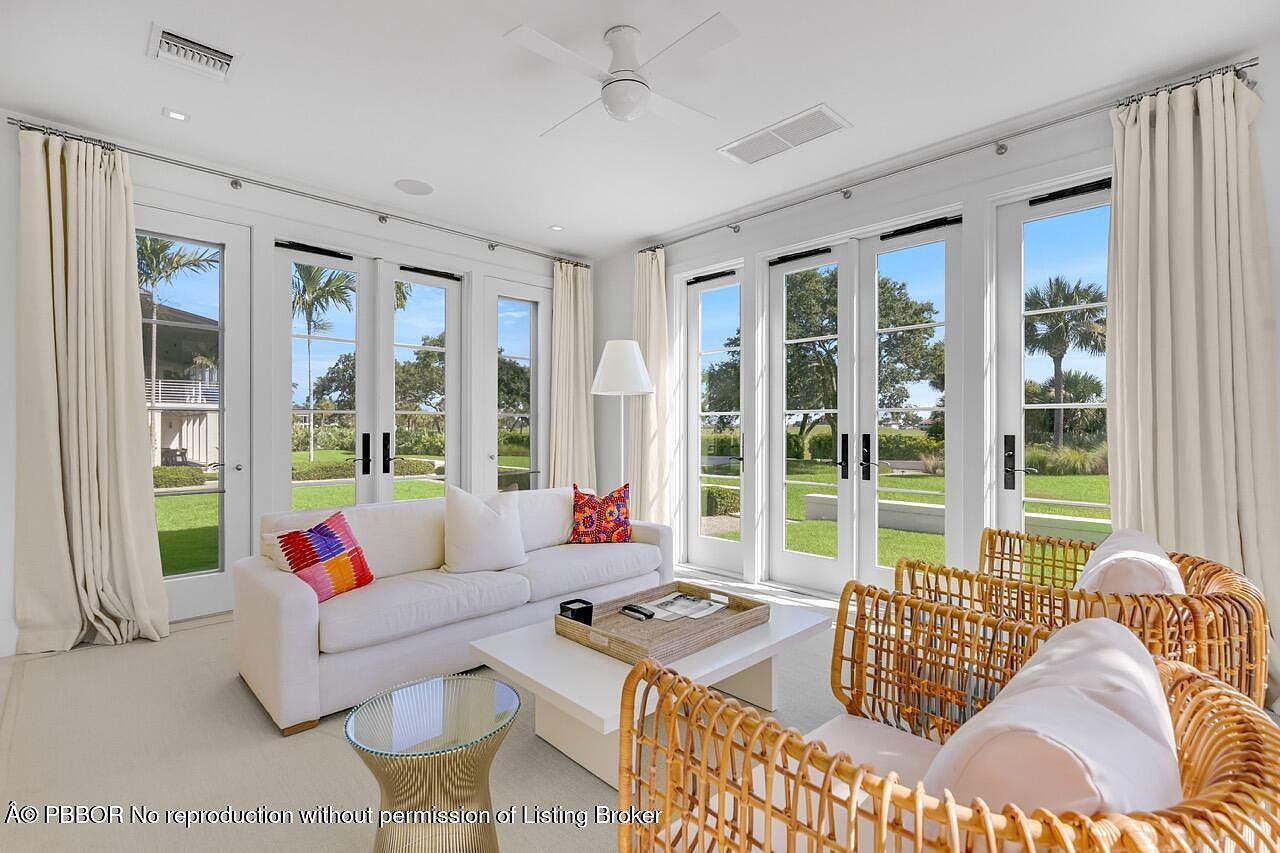 This is an interior shot of a bright and airy living room. The room features white walls, multiple French doors leading to a well-manicured lawn, and neutral-toned furniture including a white sofa with colorful pillows and wicker chairs. The overall aesthetic is clean, modern, and inviting, emphasizing natural light and outdoor views.
