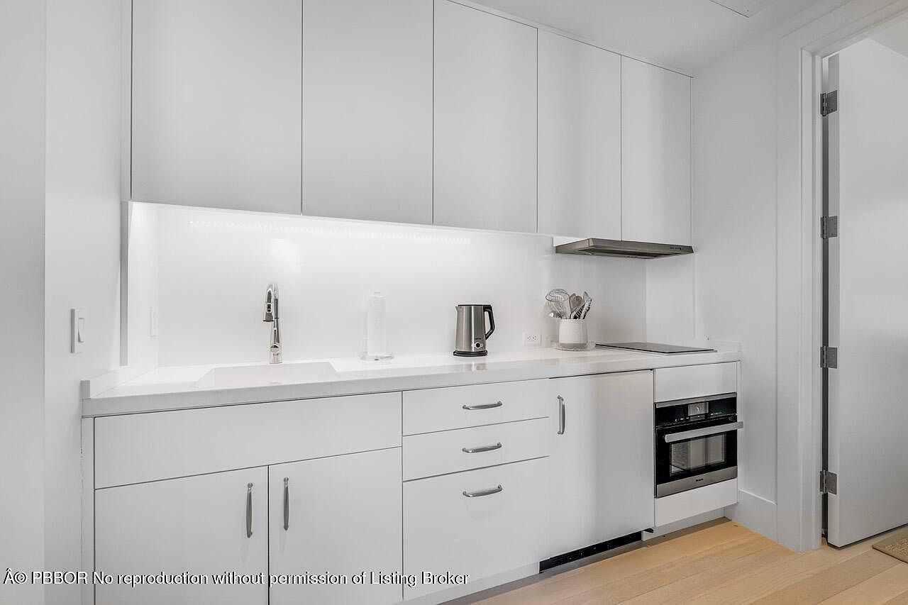 This is a well-lit, modern kitchen featuring sleek white cabinetry and countertops. The kitchen includes a stainless steel kettle, a built-in oven, and a minimalist sink with a modern faucet. The overall impression is clean, bright, and functional, ideal for a contemporary living space.