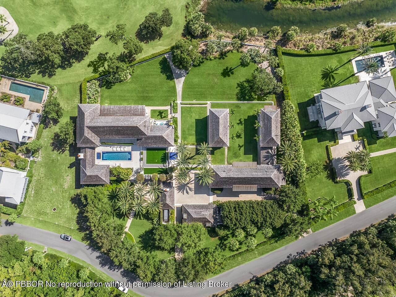 This is an aerial view of a luxurious estate showcasing multiple structures with dark wood-shingled roofs, surrounded by lush green lawns and mature trees. A large swimming pool and meticulously landscaped gardens add to the property's appeal. The overall impression is one of opulence and privacy.