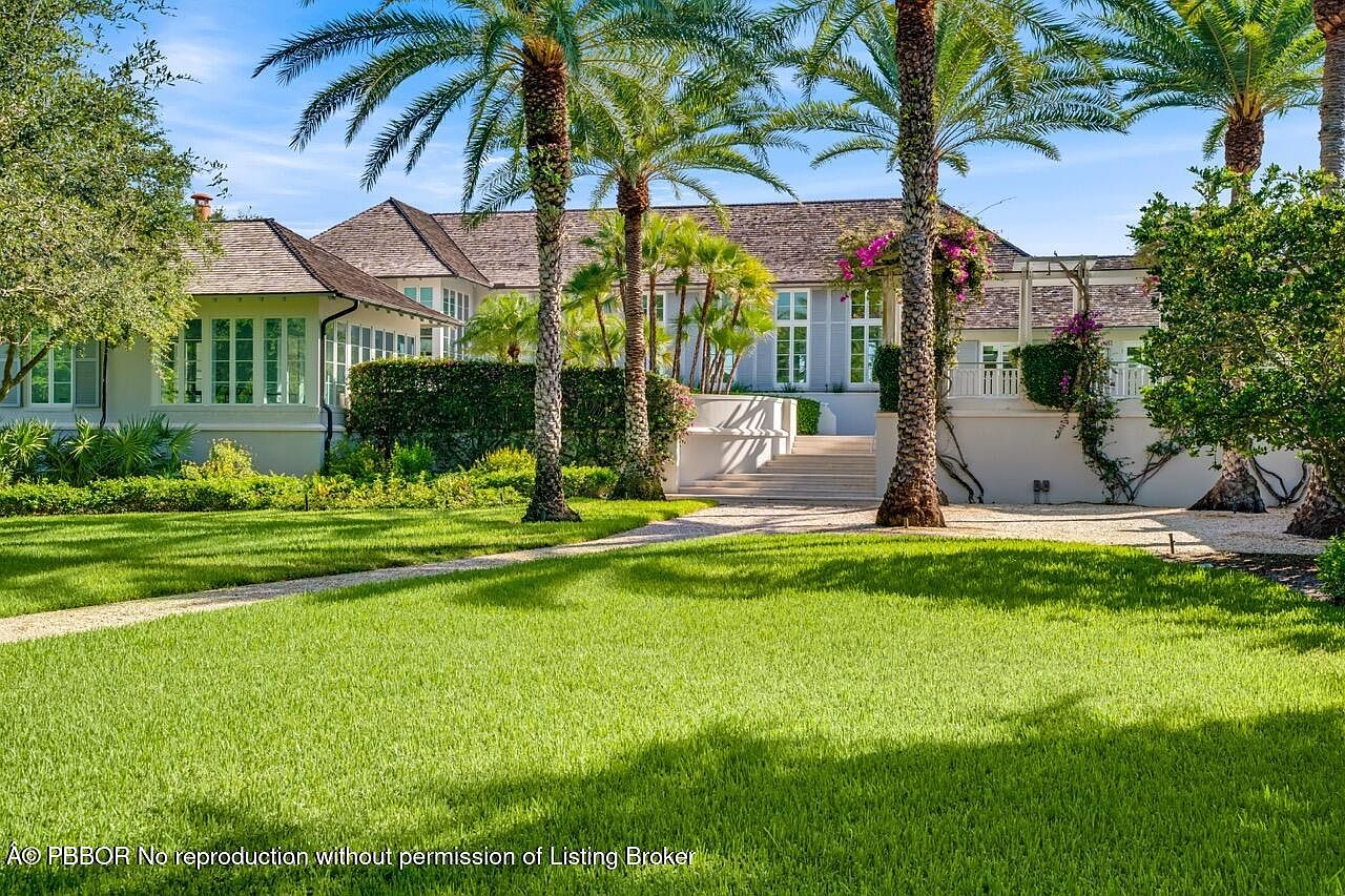 This image showcases the front exterior of a luxurious home, framed by lush green lawns and mature palm trees. The house features a light-colored facade, a multi-level roofline, and elegant architectural details, including a grand entrance with steps leading up to the main level. The landscaping is meticulously maintained, enhancing the property's curb appeal and creating a serene, inviting atmosphere.