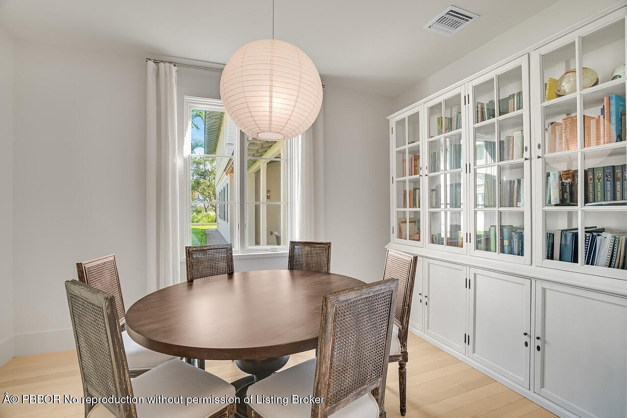 This is an interior shot of a dining room featuring a round wooden table surrounded by four chairs with woven backs and light-colored cushions. A large, round paper lantern hangs above the table, providing soft lighting. A built-in white cabinet with glass doors displays books and decorative items, adding a touch of elegance and storage to the space. The room is bright and airy, with natural light streaming in through a window.