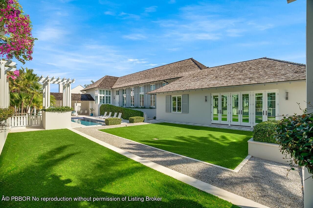 This is a beautifully landscaped backyard featuring a well-manicured lawn, a gravel pathway, and a swimming pool. The house has a light-colored exterior with a dark roof and multiple windows. The overall impression is one of luxury and tranquility, perfect for outdoor relaxation and entertainment.