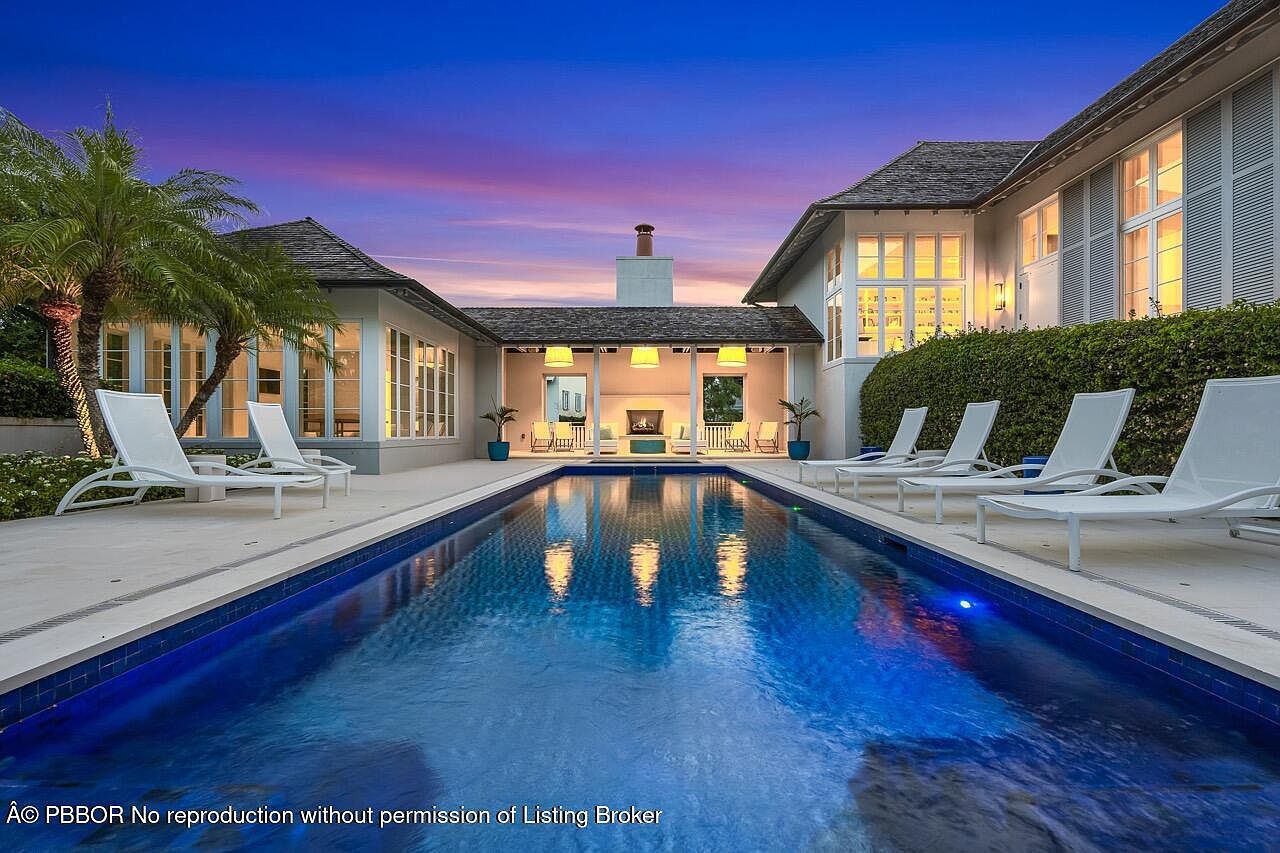 This image showcases a luxurious backyard pool area at dusk. The pool is surrounded by lounge chairs and lush landscaping, with a covered outdoor living space featuring a fireplace in the background. The overall impression is one of relaxation and high-end outdoor living.