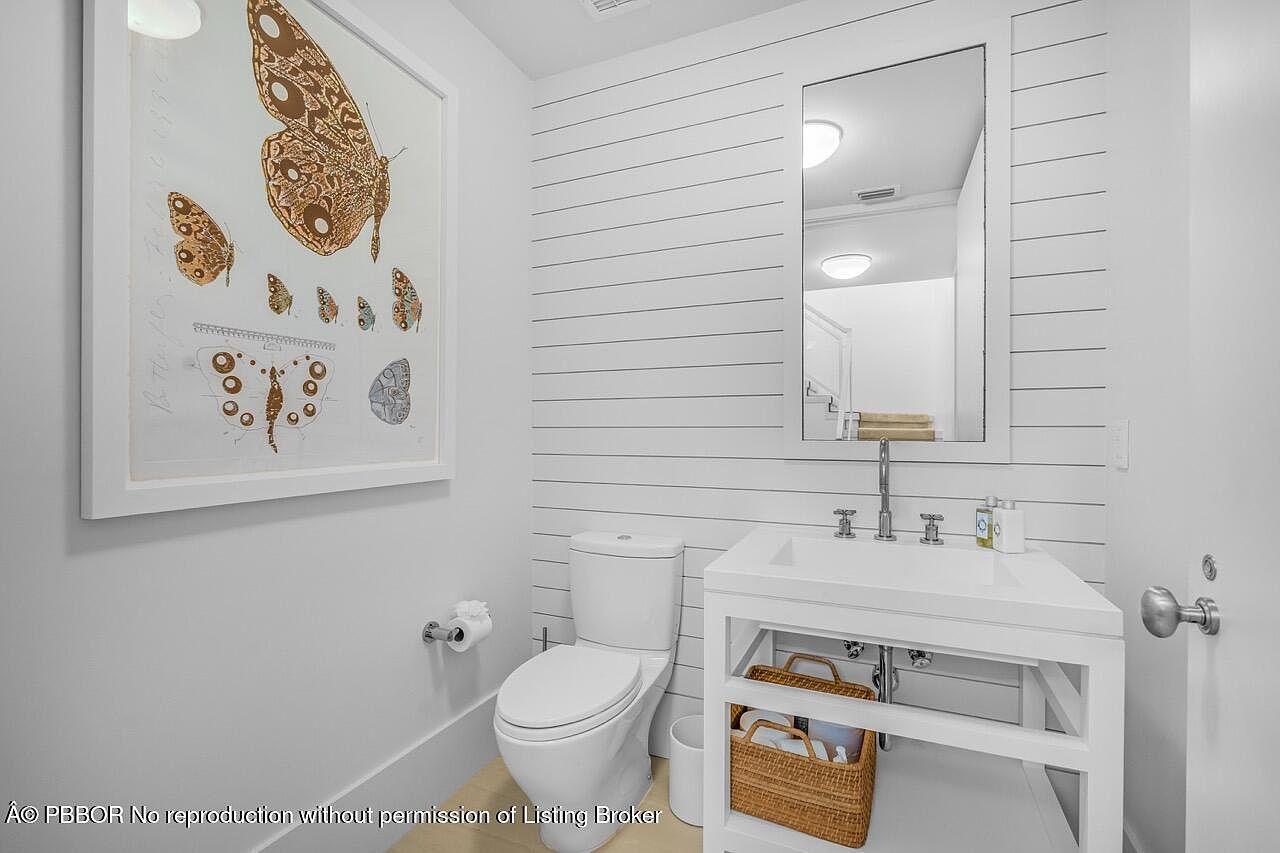 This is a well-lit guest bathroom featuring a white vanity with a sink, a toilet, and a framed mirror. The walls are decorated with horizontal shiplap and a framed butterfly print, adding a touch of elegance. A woven basket under the sink provides storage and complements the clean, modern aesthetic.