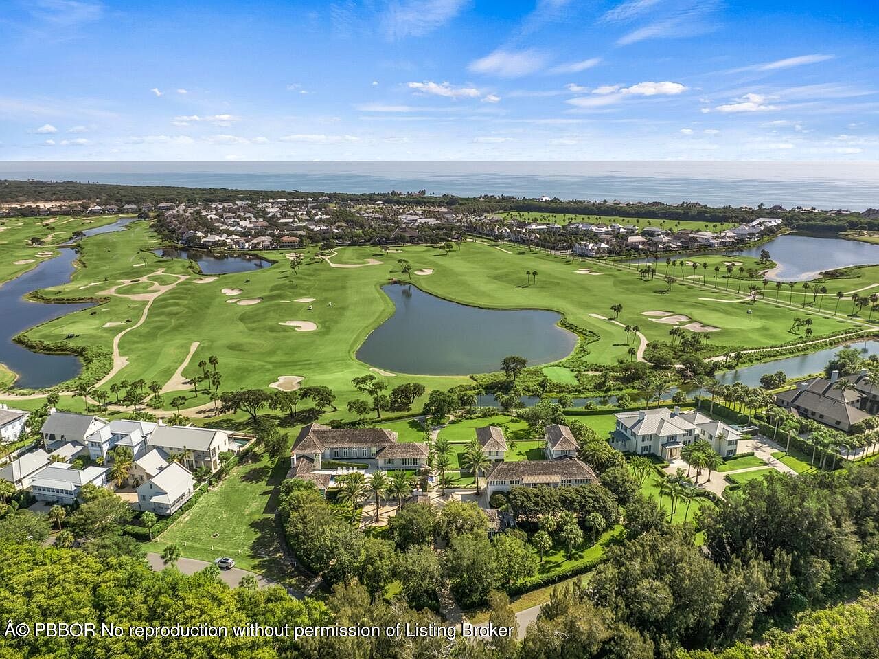 This aerial shot showcases a luxurious estate nestled within a meticulously maintained golf course community. The property features multiple buildings with elegant architecture, surrounded by lush landscaping and mature trees. The view extends to the ocean in the distance, highlighting the prime location and upscale lifestyle.