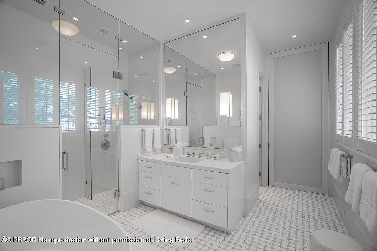 This is a bright and modern primary bathroom featuring a glass-enclosed shower, a double vanity with white cabinetry and countertops, and a large mirror. The floor is tiled with a small, circular pattern, and natural light streams in through shuttered windows. The overall impression is clean, luxurious, and well-lit.