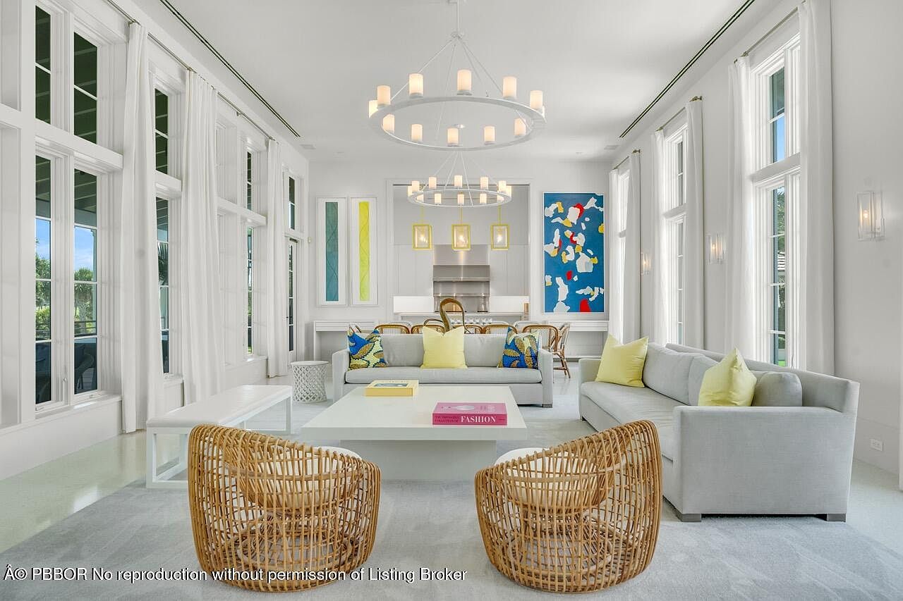 This is a bright and airy living room featuring large windows with white curtains, allowing ample natural light. The room is furnished with two light gray sofas, two rattan chairs, and a white coffee table, creating a comfortable and inviting space. The decor includes colorful artwork and a modern chandelier, adding a touch of elegance and sophistication.