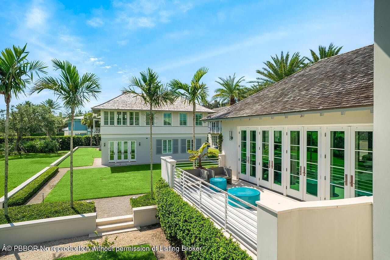 This image showcases a beautifully landscaped patio or deck area of a luxury home. The space features modern outdoor furniture, including seating and vibrant blue accent tables, set against a backdrop of lush greenery and palm trees. The architectural style includes white railings and large glass doors, suggesting a seamless transition between indoor and outdoor living spaces, enhancing the property's appeal.