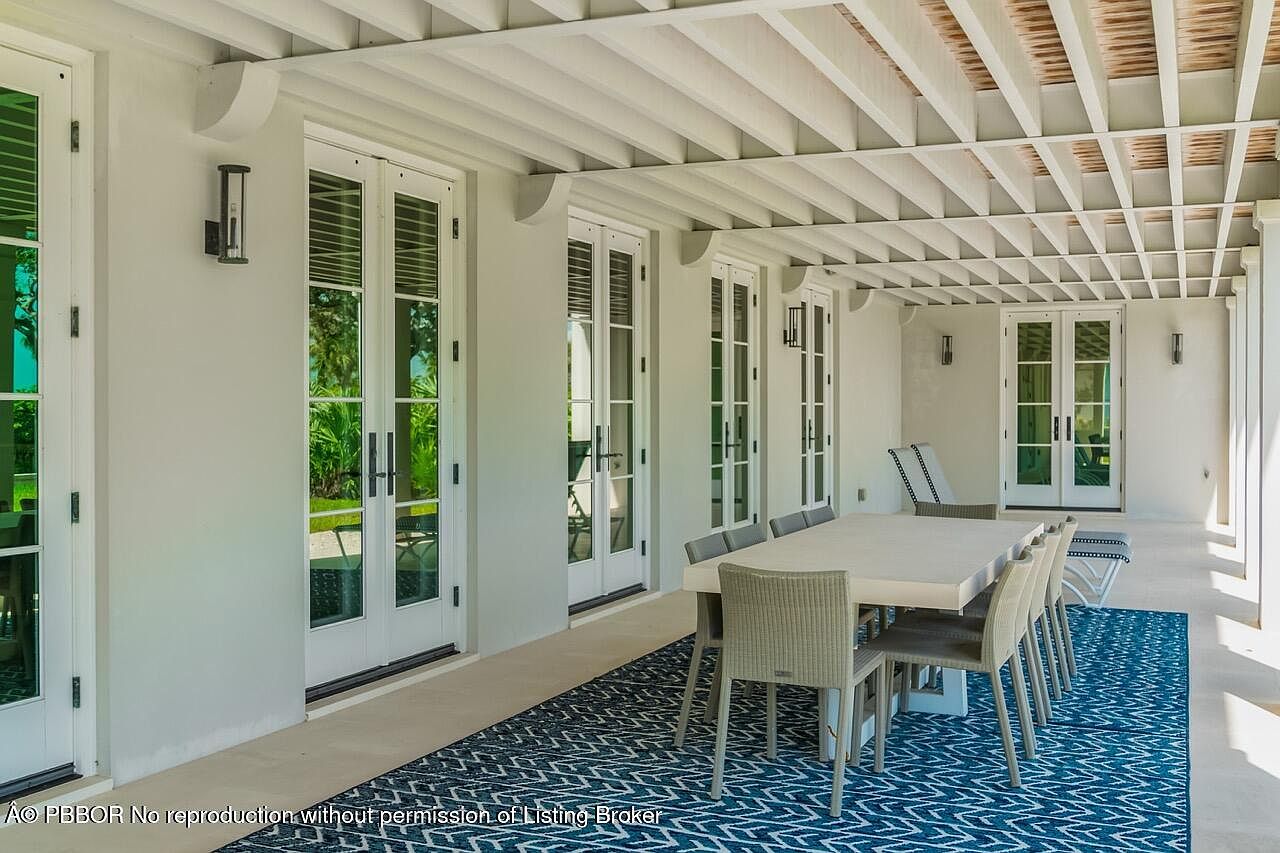 This is a well-appointed outdoor patio area, featuring a long dining table with seating for eight, set upon a blue patterned rug. The patio is covered by a white pergola-style roof, and several sets of French doors lead into the interior of the home. The space is designed for outdoor dining and relaxation, offering a seamless transition between indoor and outdoor living.