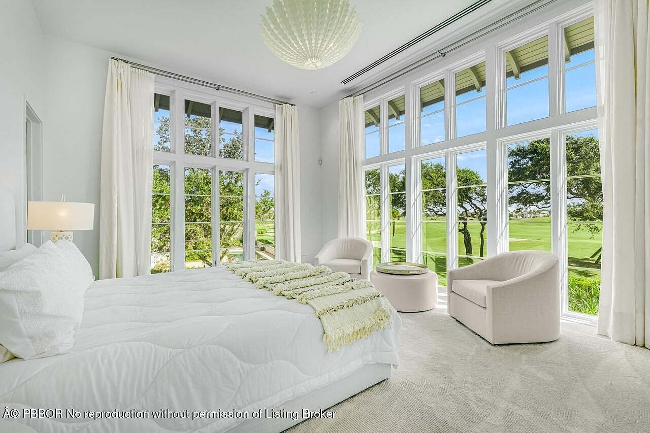 This is a bright and airy primary bedroom featuring large windows that offer a view of a golf course. The room is decorated in a neutral color palette with white bedding, curtains, and furniture. The overall impression is one of luxury and tranquility, perfect for relaxation.