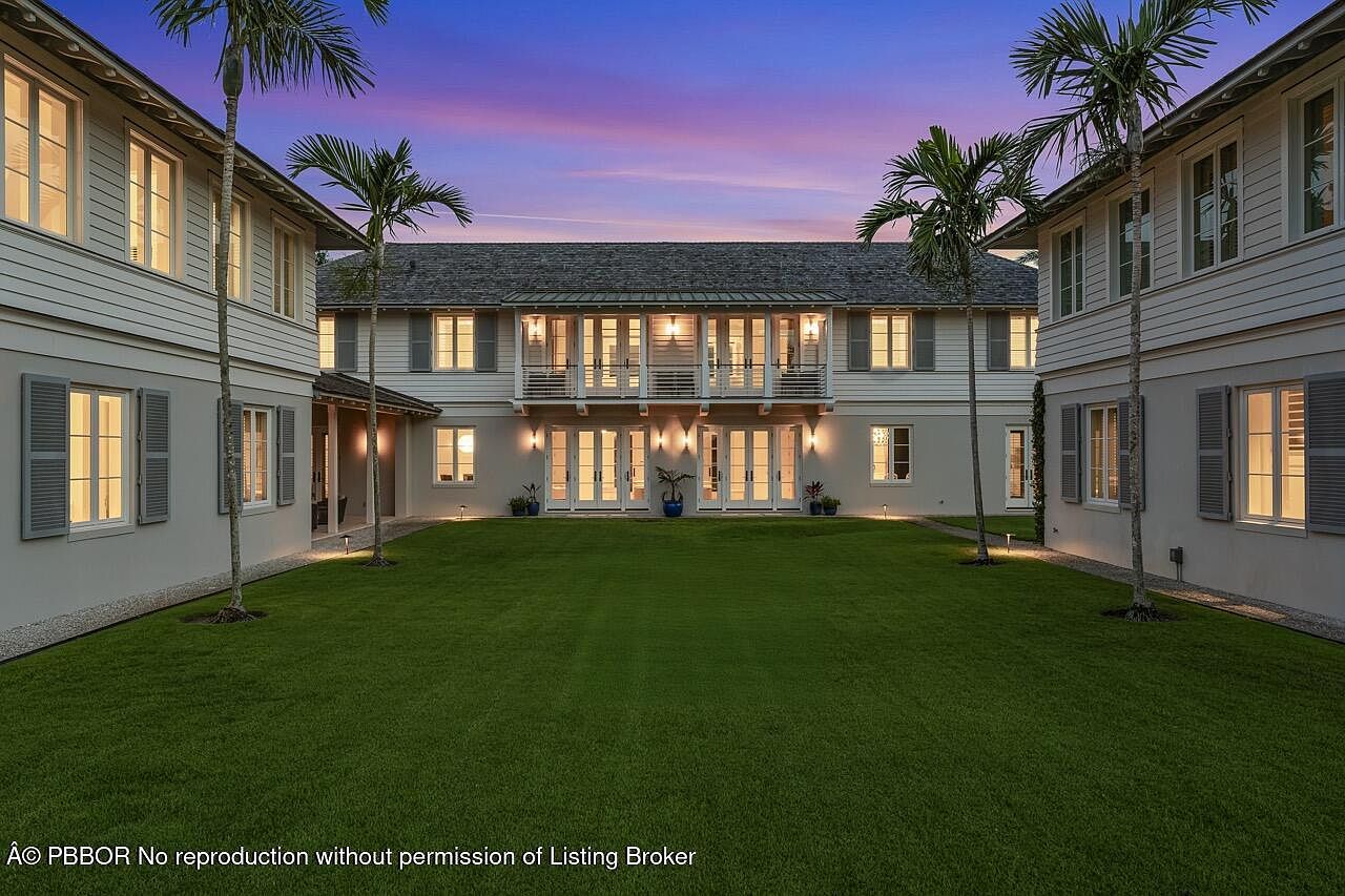 This is a front exterior view of a two-story residential building with a symmetrical design. The building features a well-manicured lawn, palm trees, and multiple windows with shutters. The architecture suggests a luxurious and inviting atmosphere, perfect for a family home.