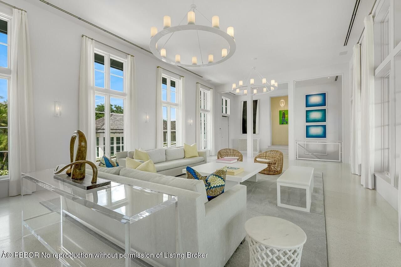 This is a bright and airy living room featuring white walls, large windows with sheer curtains, and modern lighting fixtures. The room is furnished with white sofas, decorative pillows, and a clear acrylic console table. The overall impression is clean, elegant, and spacious, perfect for entertaining or relaxing.