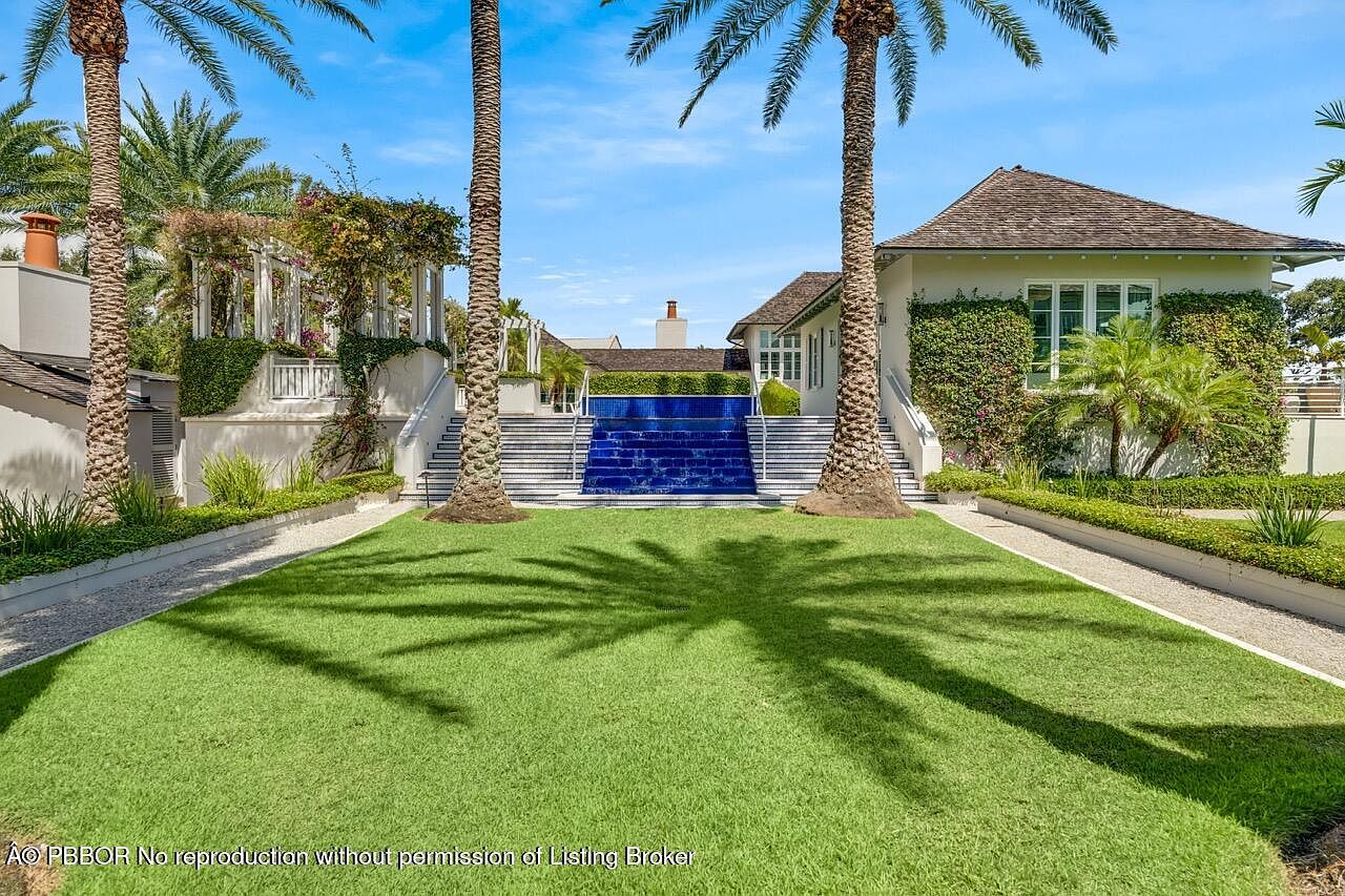 This image showcases a beautifully landscaped yard with a tiered water feature and symmetrical design. Palm trees frame the view, casting shadows on the manicured lawn. The property features elegant architectural details, including white staircases and lush greenery, creating a luxurious and serene outdoor space.