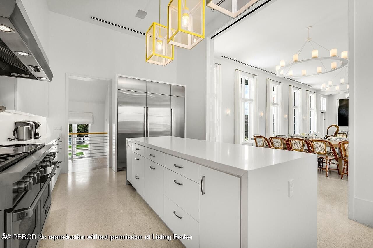 This is a bright and spacious kitchen featuring a large white island with ample storage, stainless steel appliances, and modern yellow pendant lighting. The kitchen seamlessly transitions into a dining area with a large chandelier and multiple windows, creating an open and airy atmosphere. The overall design is clean, contemporary, and inviting.