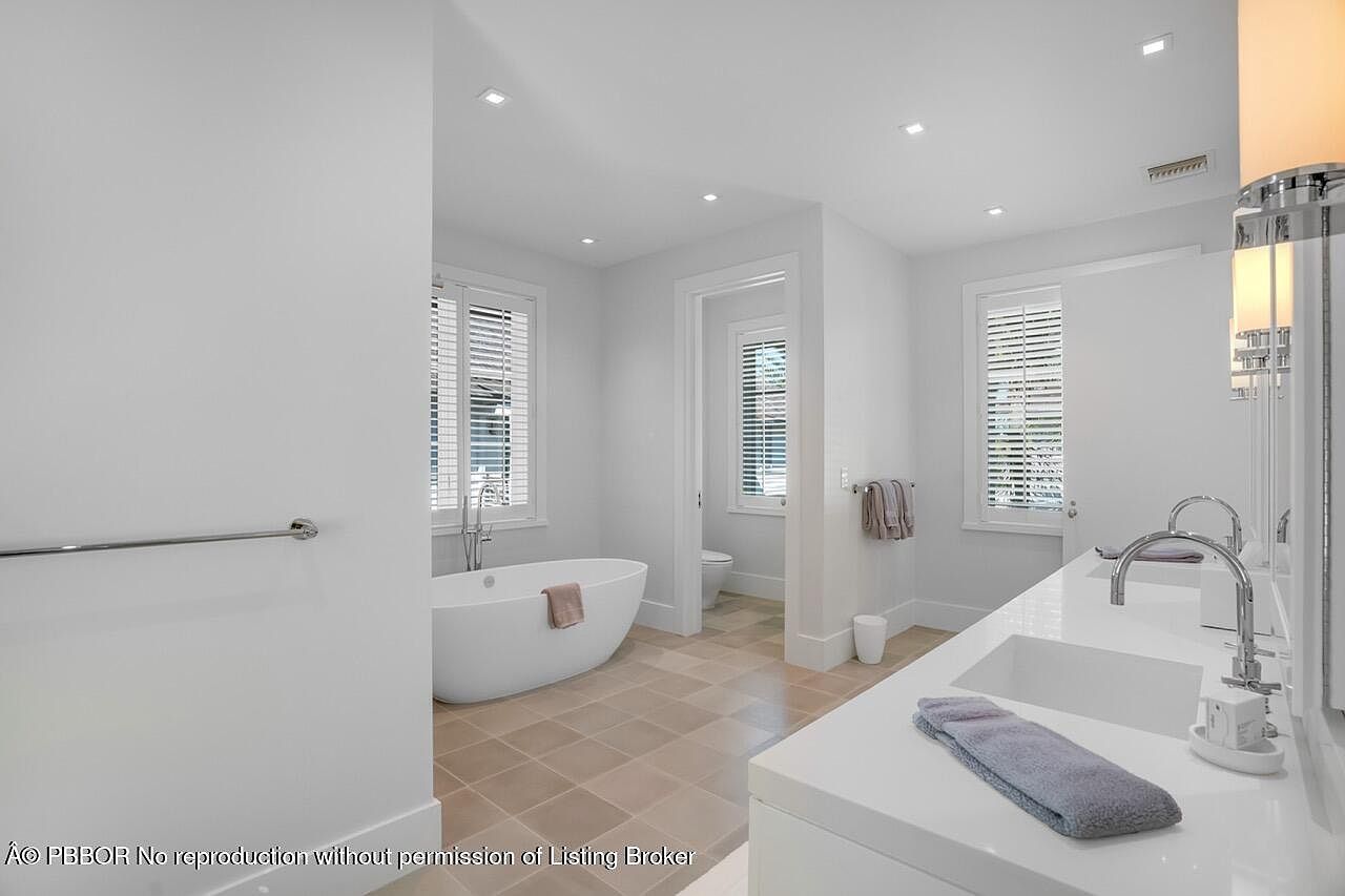 This is a bright and modern primary bathroom featuring a freestanding bathtub next to a window with blinds, a double sink vanity with white countertops, and neutral-toned tile flooring. The bathroom has a clean and minimalist design with recessed lighting and chrome fixtures, creating a luxurious and serene atmosphere. A doorway leads to a separate toilet room.