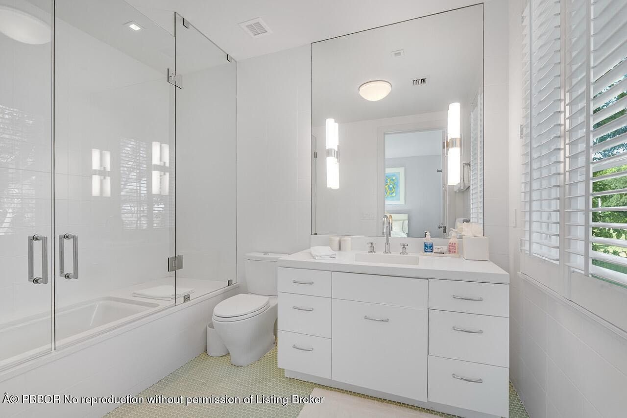This is a bright and modern bathroom featuring a glass-enclosed bathtub and shower combination. The vanity has a white countertop with ample storage space in the drawers and cabinets. A large mirror reflects the light, and the room is well-lit with both overhead and sconce lighting, creating a clean and inviting space.