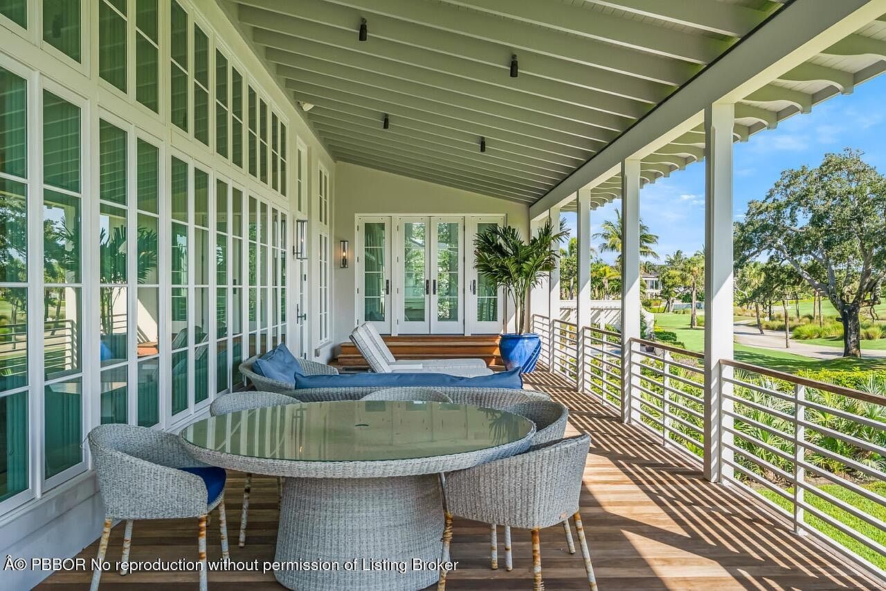 This image showcases a luxurious outdoor patio or deck area, featuring stylish wicker furniture including a round table and chairs, complemented by a comfortable lounge area with blue cushions. The space is partially covered, offering shade, and overlooks a beautifully manicured lawn and golf course, creating a serene and upscale ambiance. Large windows and doors provide seamless indoor-outdoor living.