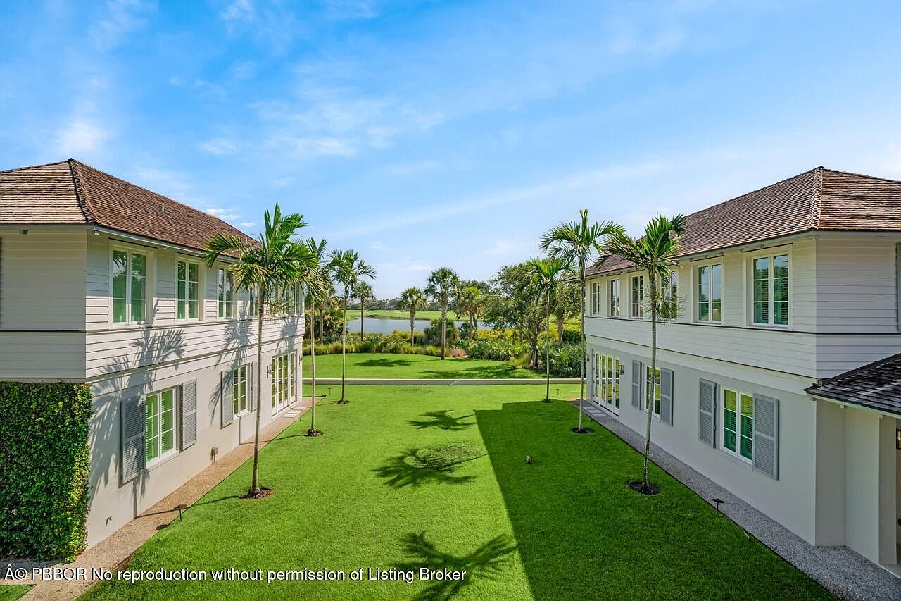 This image showcases a meticulously maintained lawn situated between two elegant, two-story homes. The lush green grass is punctuated by young palm trees, leading the eye towards a serene lake and distant trees under a bright blue sky. The scene evokes a sense of luxury, tranquility, and spaciousness, highlighting the property's desirable outdoor living space.