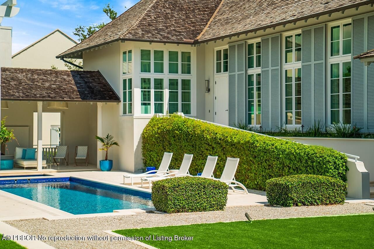 This image showcases a luxurious backyard featuring a pristine pool with a light-colored stone surround. The house has a light exterior with blue shutters and large windows, complemented by meticulously maintained greenery and manicured bushes. The scene exudes a sense of tranquility and upscale outdoor living.